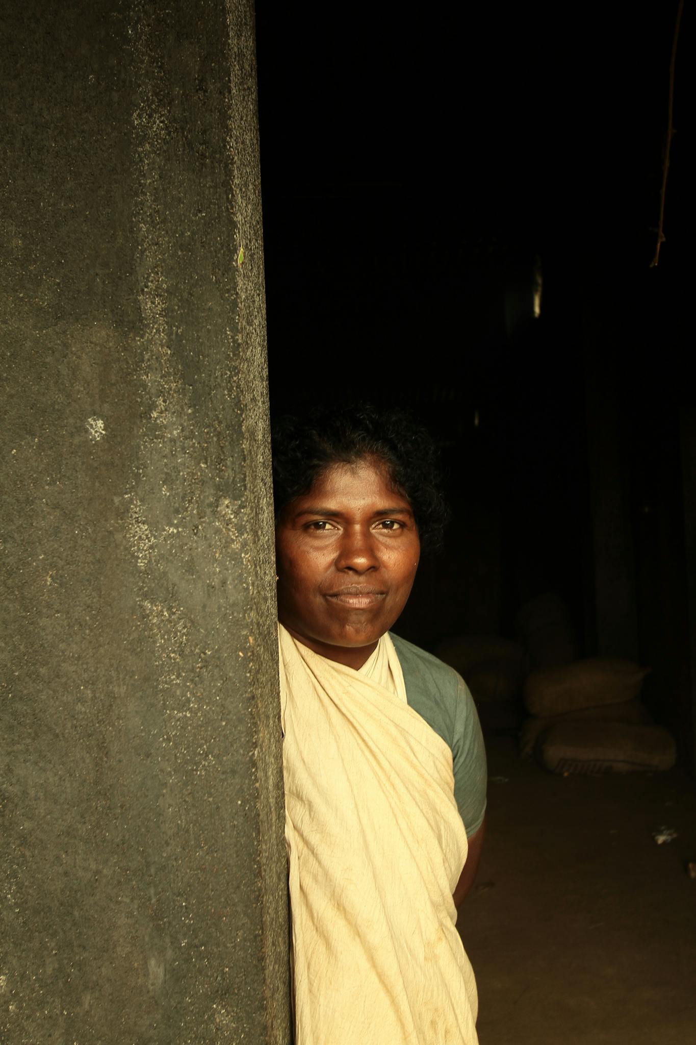 A woman in a yellow sari stands by a concrete wall, looking confidently at the camera.